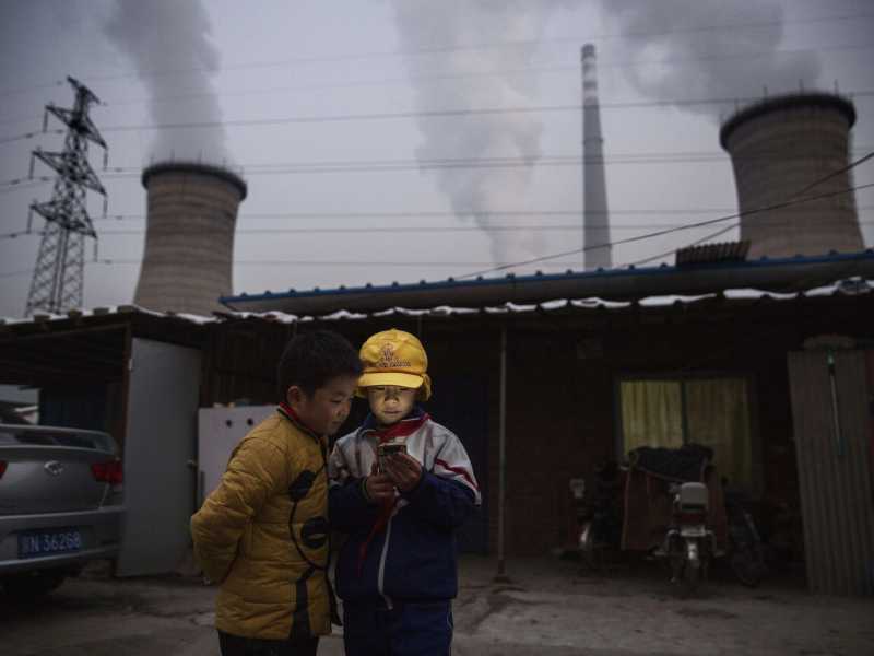 Ragazzi cinesi guardano lo smartphone davanti alla loro casa accanto a una centrale a carbone alla periferia di Pechino (Foto di Kevin Frayer/Getty Images)&nbsp;<br />