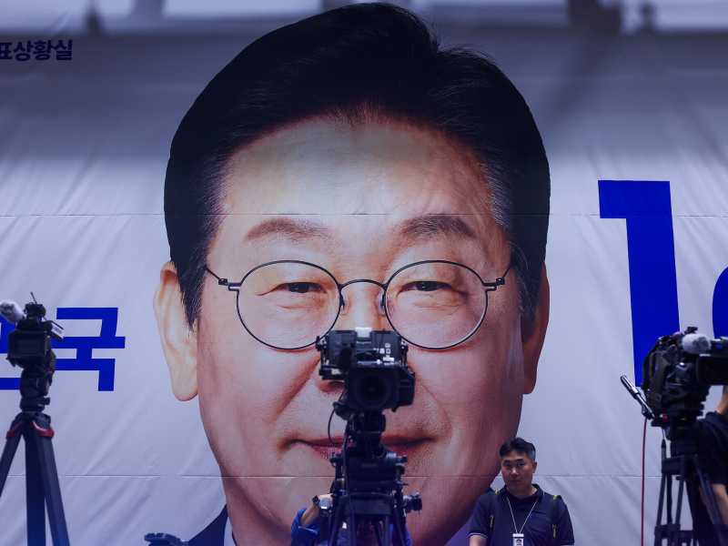 I cameraman sistemano le loro attrezzature davanti a uno striscione con il volto del candidato presidenziale Lee Jae-myung (Daniel Ceng/Getty Images)<br />