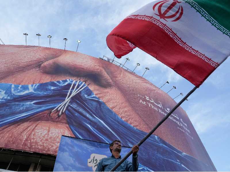 Un uomo sventola una bandiera iraniana sotto un cartellone con l'immagine dello Stretto di Hormuz e delle labbra cucite a Trump, in una piazza del centro di Teheran (AP/LaPresse)